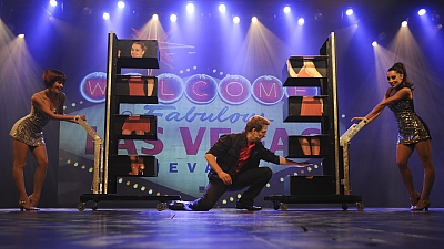 Ein Magier führt auf einer Bühne mit zwei Assistentinnen einen Zaubertrick auf.