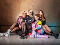 Vier junge Frauen in 90er Jahre Look sitzen lässig vor einer Wand.