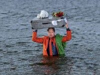 Ein orange gekleideter Mann steht bis zur Brust im Wasser und hält einen überquellenden Reisekoffer über dem Kopf.