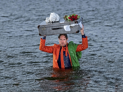 Patrick Nederkoorn Ein orange gekleideter Mann steht bis zur Brust im Wasser und hält einen überquellenden Reisekoffer über dem Kopf.