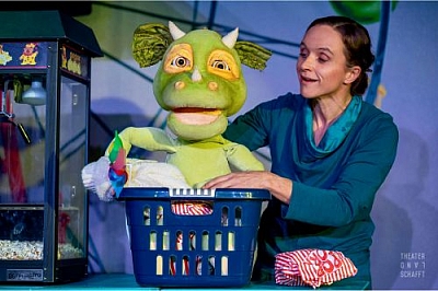 Grüne Drachen-Handpuppe in einem blauen Wäschekorb neben einer Schauspielerin die die Handpuppe bewegt.