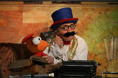 Schauspieler mit Zylinder, Schnurrbart und Brille, der ein Hörrohr auf eine Enten-Handpuppe richtet.