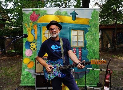 Das Bild zeigt einen Mann mit Hut vor einem bunten Hintergrund, der eine blaue Gitarre spielt. 