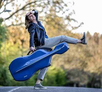 Musikerin in Lederjacke und Jeans mit blauer Cellotasche in der Hand.